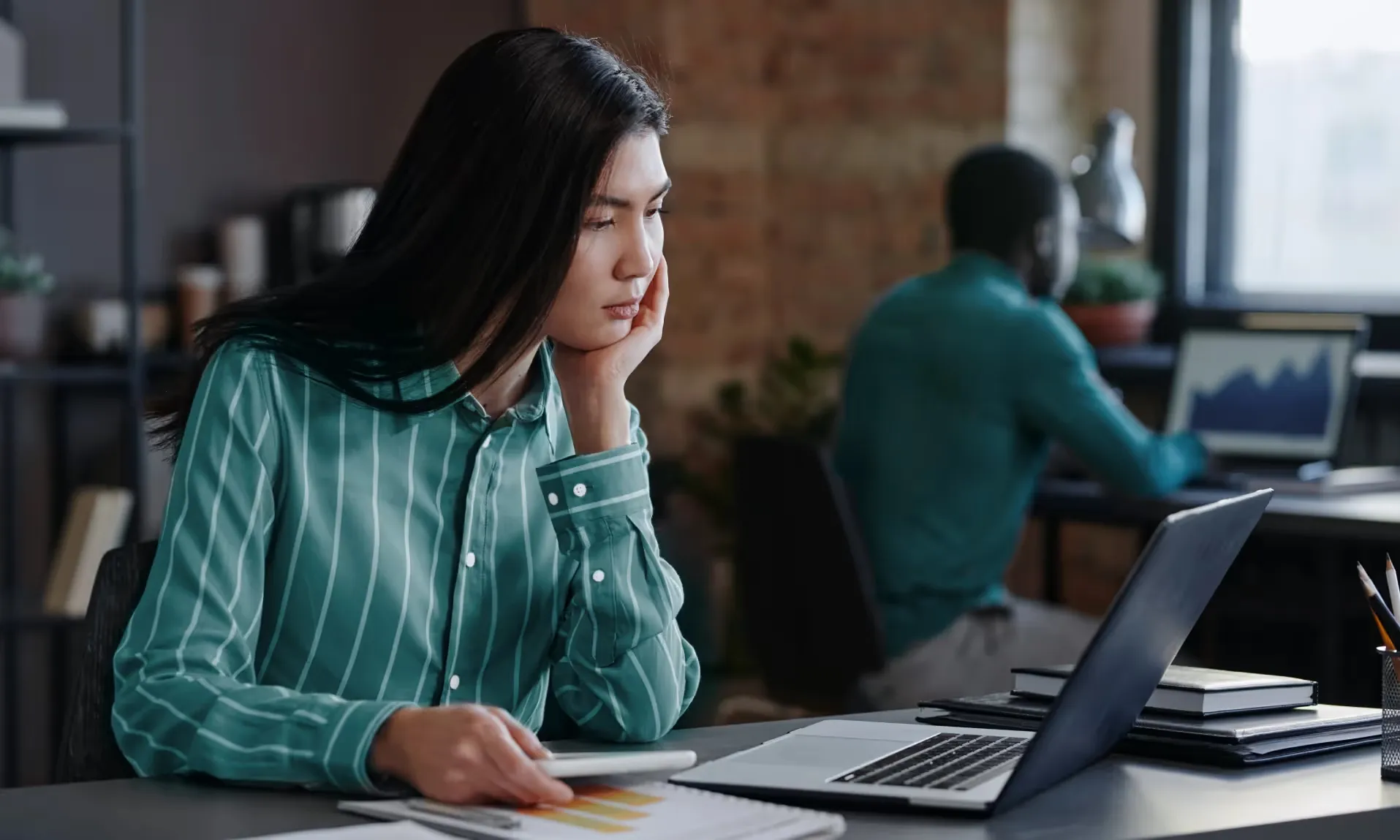 Mulher pensativa diante de um laptop, com um colega trabalhando ao fundo