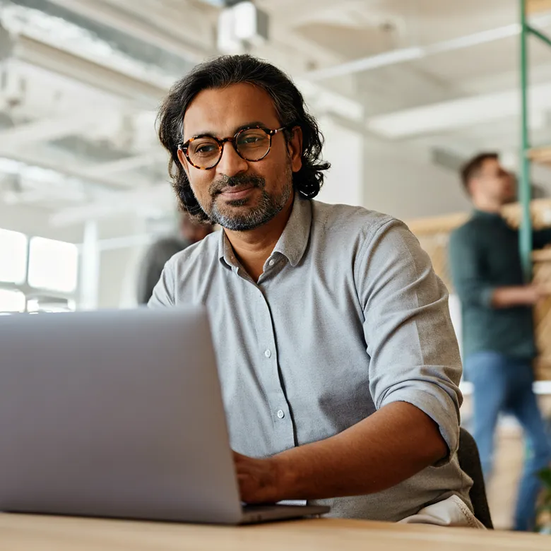 Man in glasses sitting at a desk, using a laptop in an open-plan office
