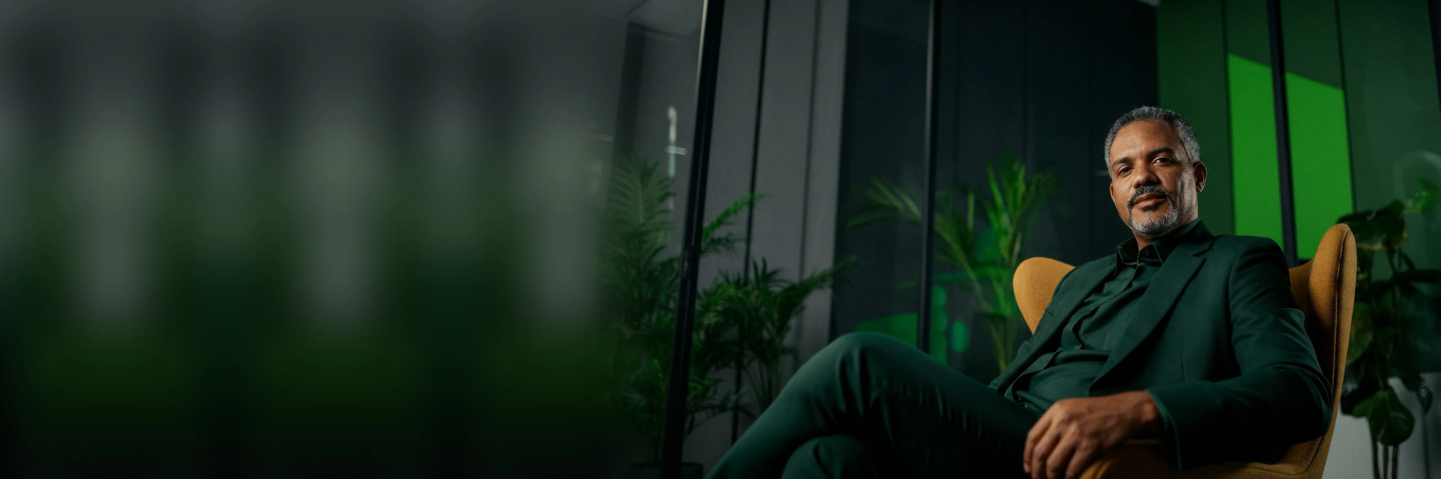 Man in a green suit seated in a yellow armchair in an office with plants.