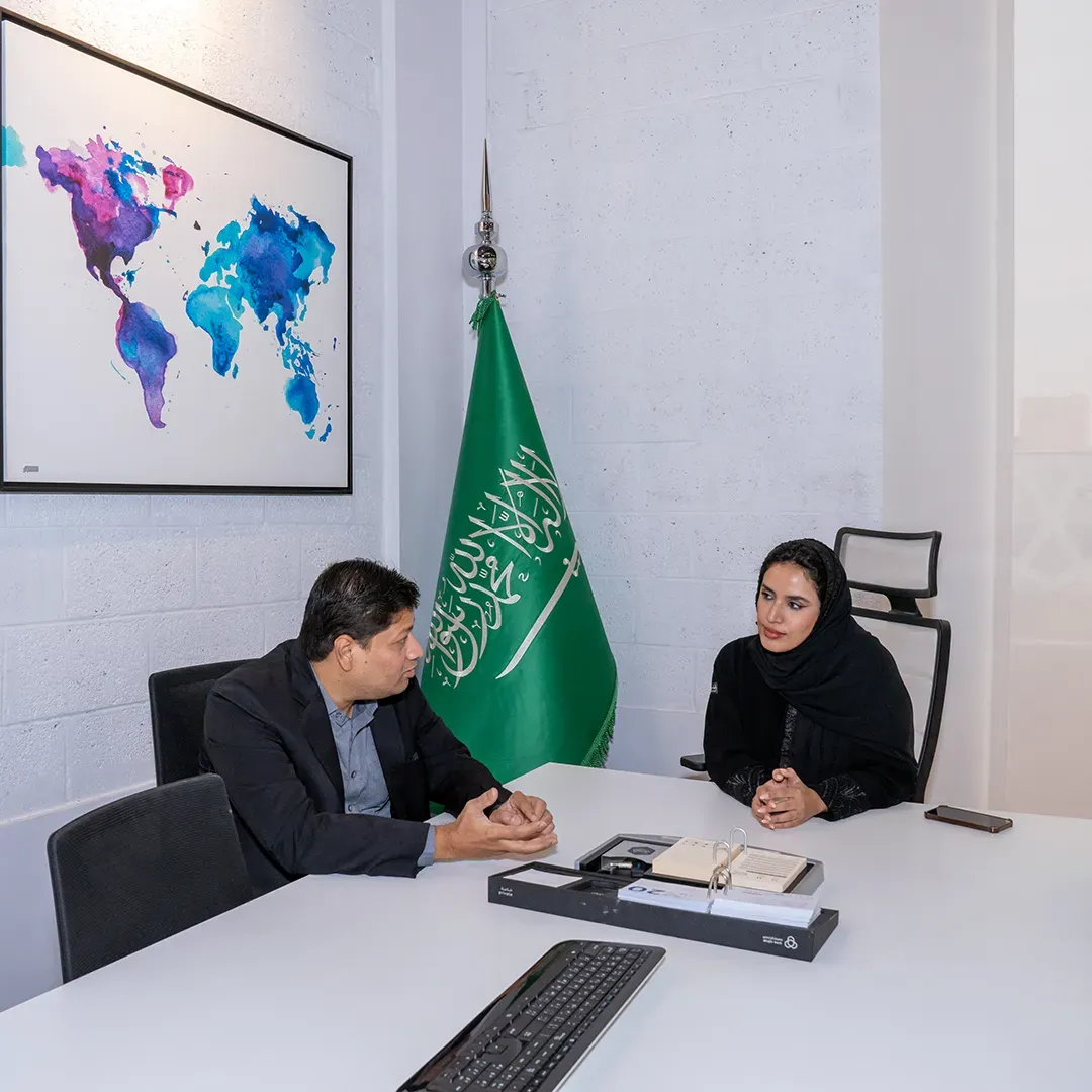 Two professionals in a meeting in a room with the Saudi Arabian flag